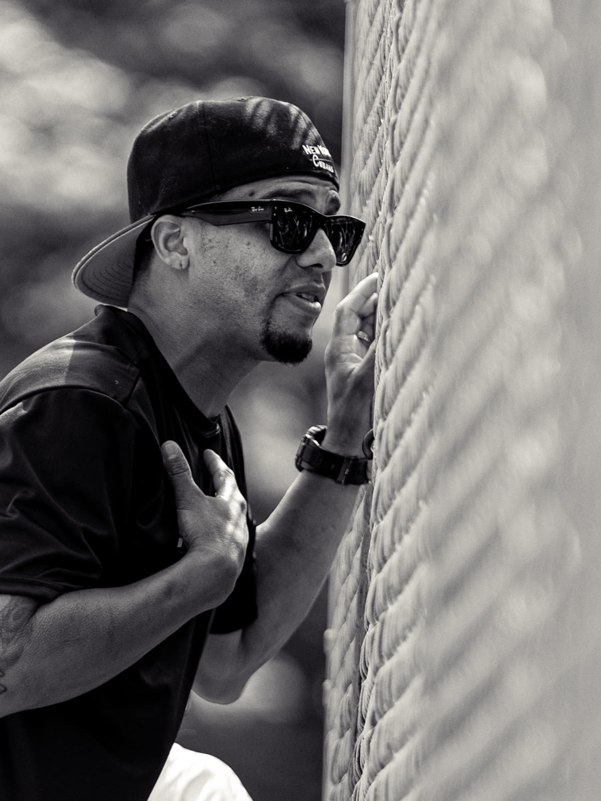 Black-and-white photograph of a man in a New York Cubans cap and sunglasses leaning against the backstop fence, one hand to his chest.