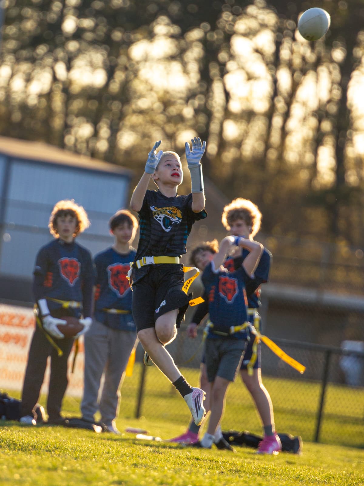 A young flag-football player in a Jaguars jersey jumps to gather a high pass in golden evening light, teammates watching from the sideline.
