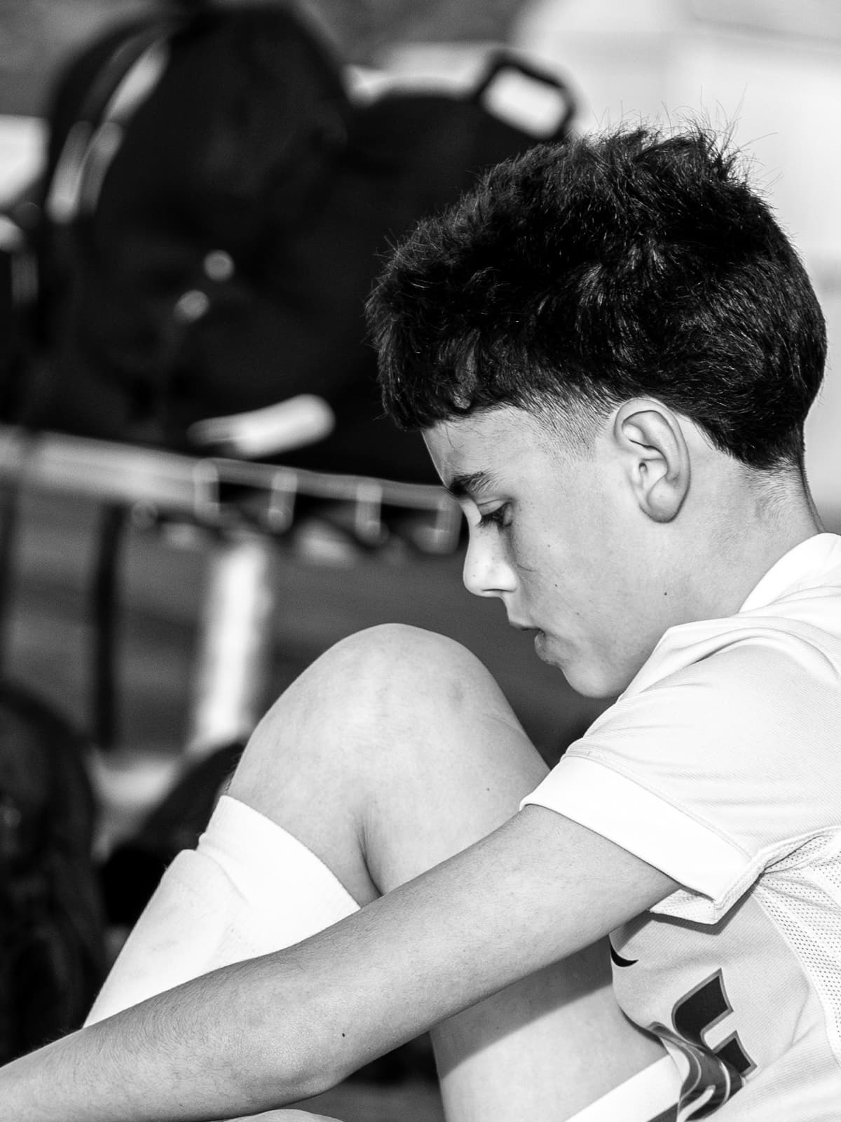 Black-and-white photograph of a teenage soccer player sitting with his knees up and head down beside the equipment rack.