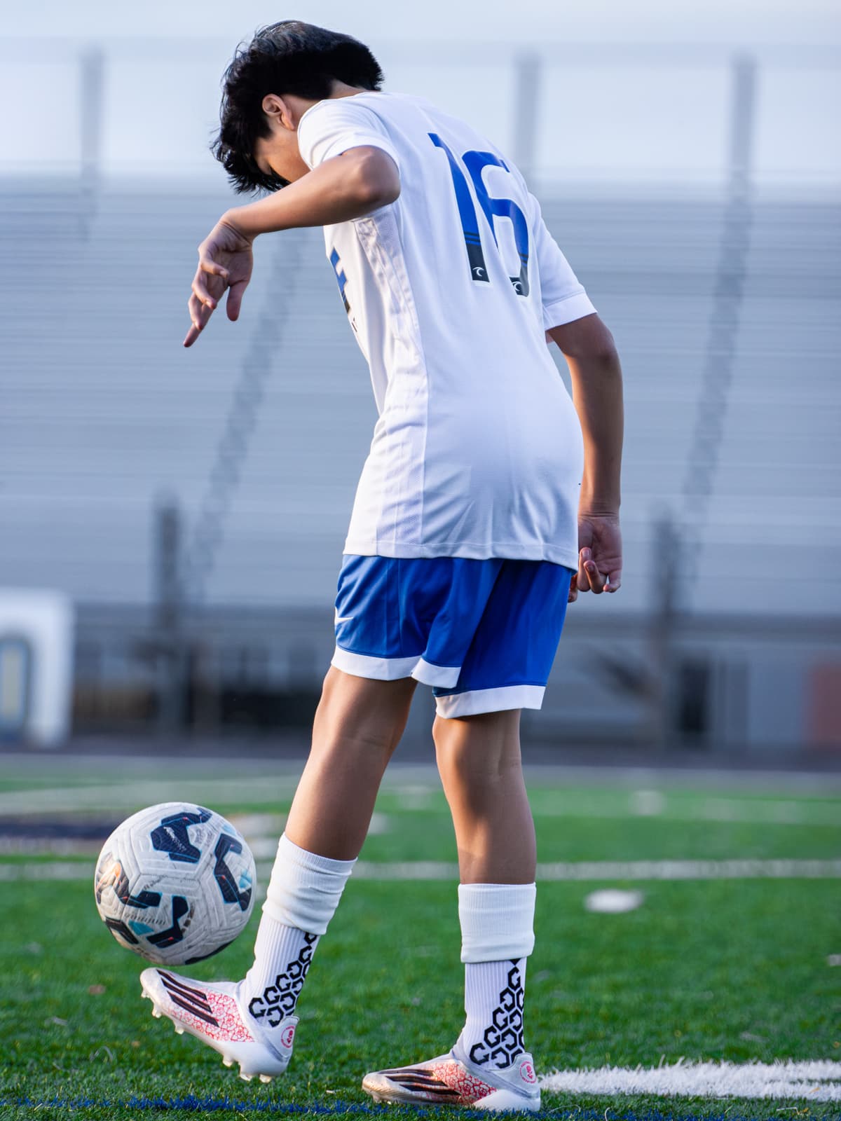 A soccer player in a white number 16 kit juggles the ball on an empty field, seen from behind against the stands.