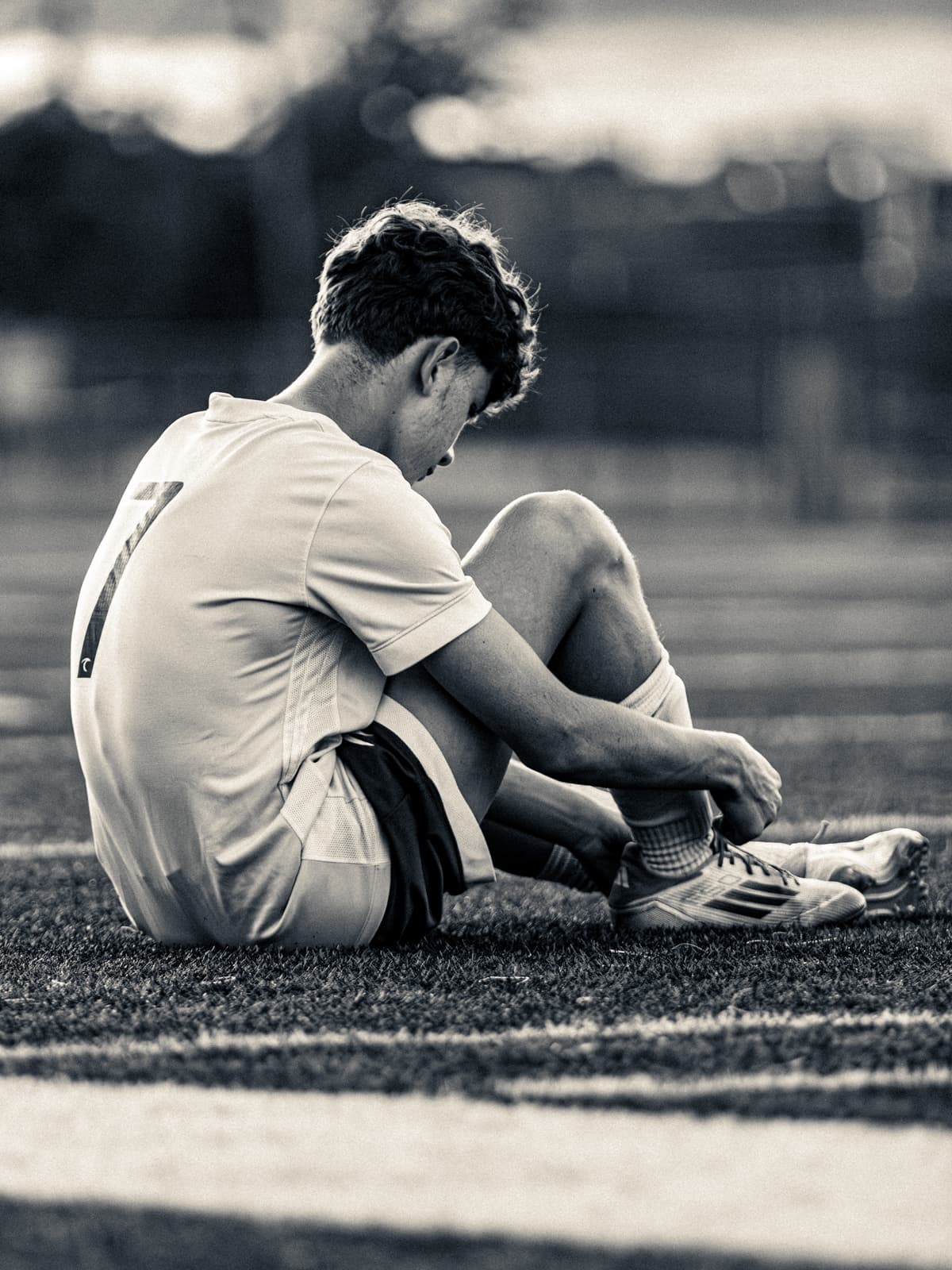 Sepia-toned photograph of a soccer player sitting on the turf to lace up a boot, head down in the last of the light.