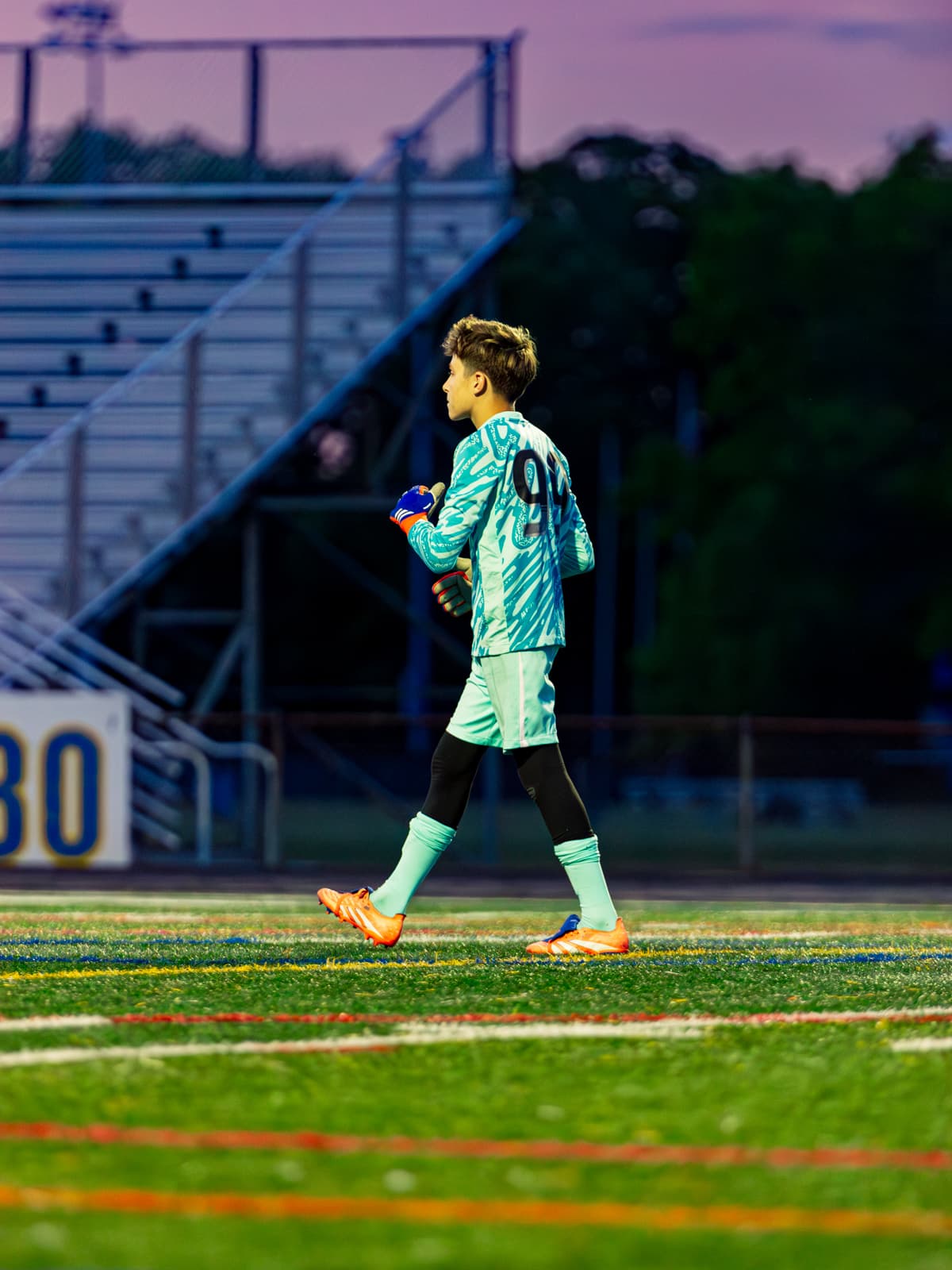 A soccer goalkeeper in a mint-green kit walks across the field at dusk under a purple sky, empty bleachers behind.
