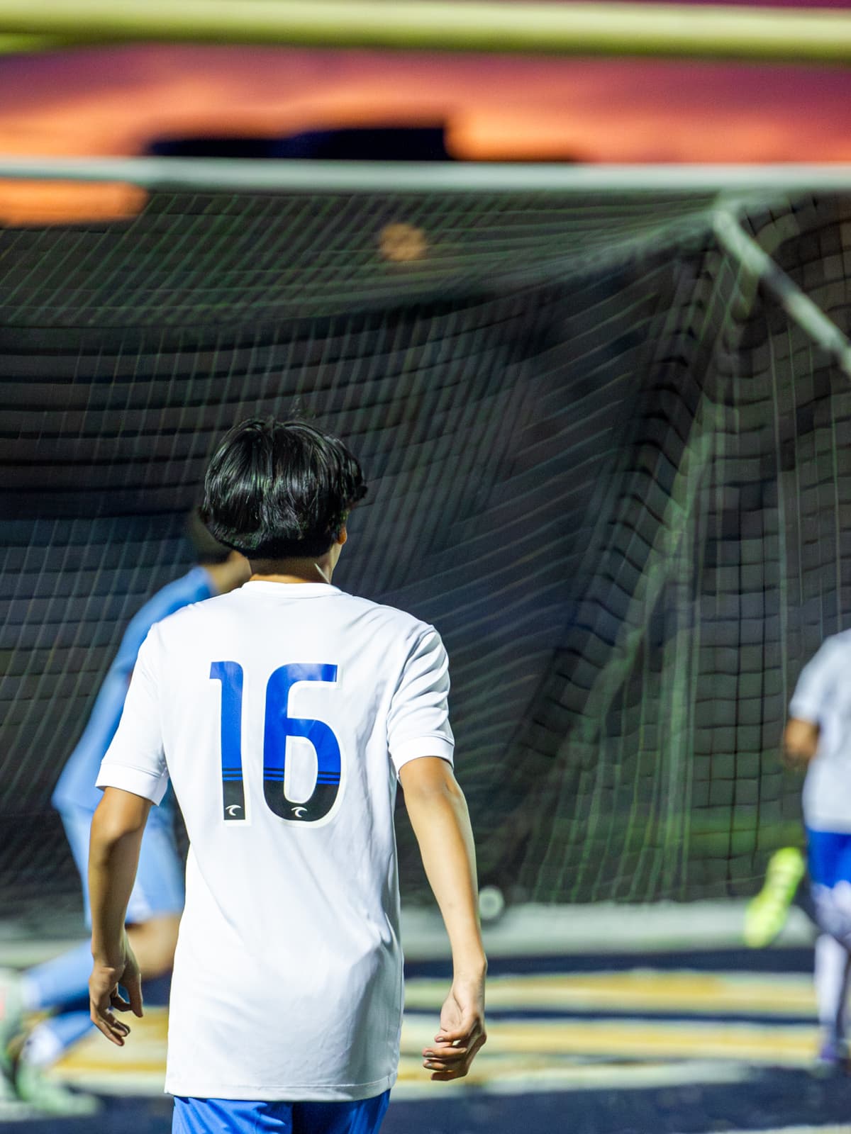 A soccer player in a white number 16 kit walks toward the goal net during a night game, seen from behind.
