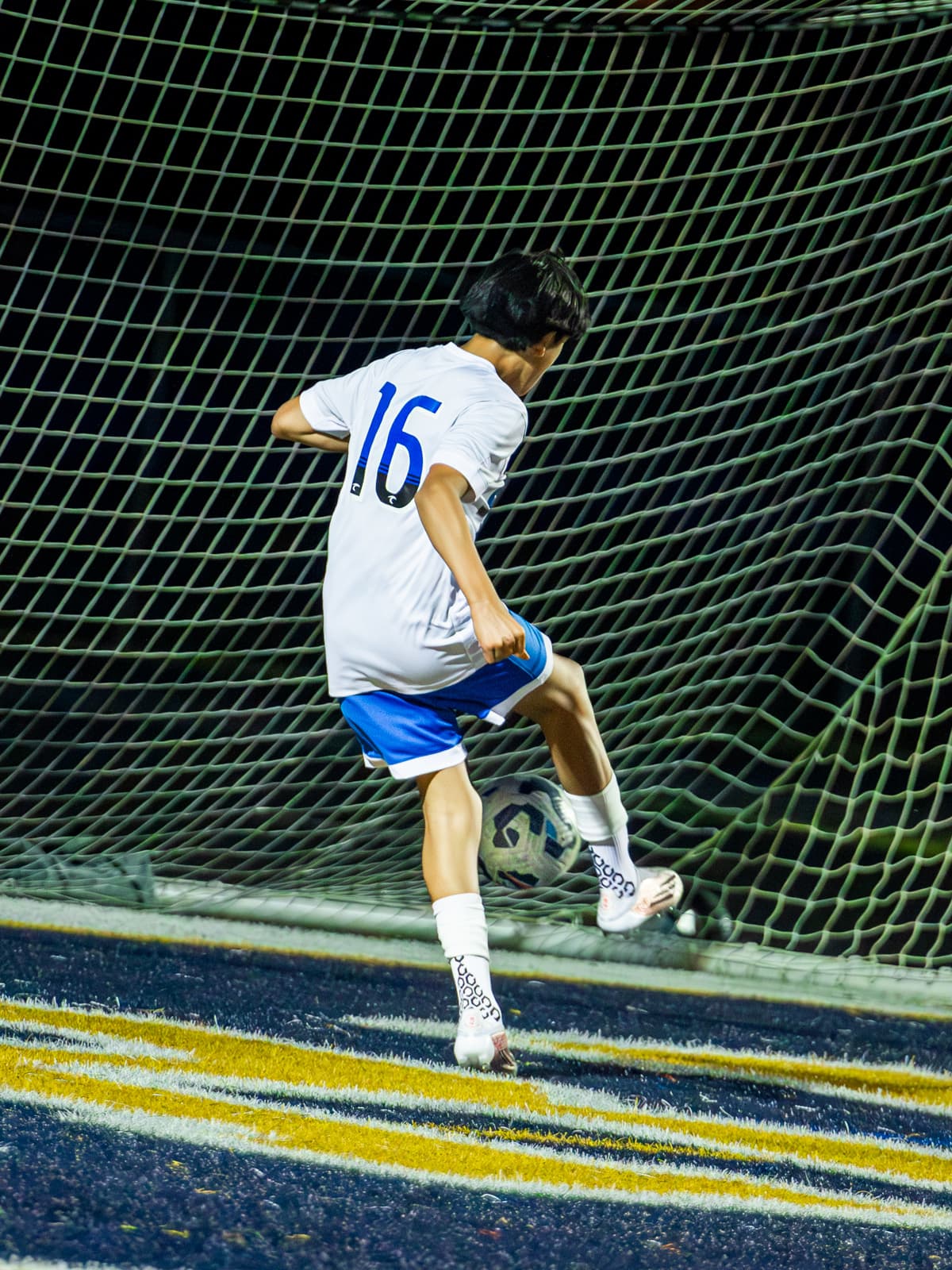 A soccer player in a white number 16 kit flicks the ball up just in front of the goal net during a night game, seen from behind.