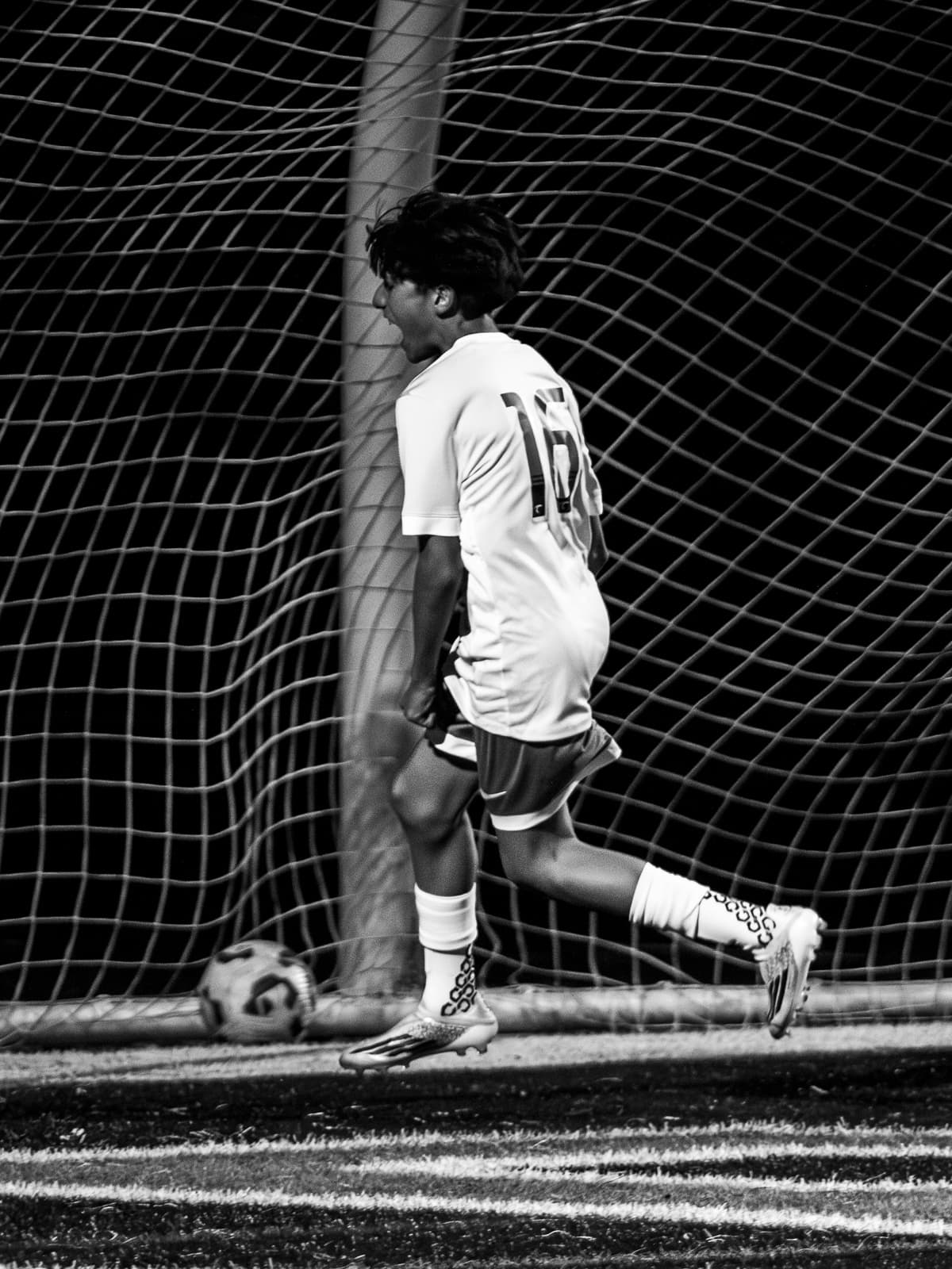 Black-and-white photograph of a soccer player wheeling away in celebration as the ball settles in the net behind him under the lights.