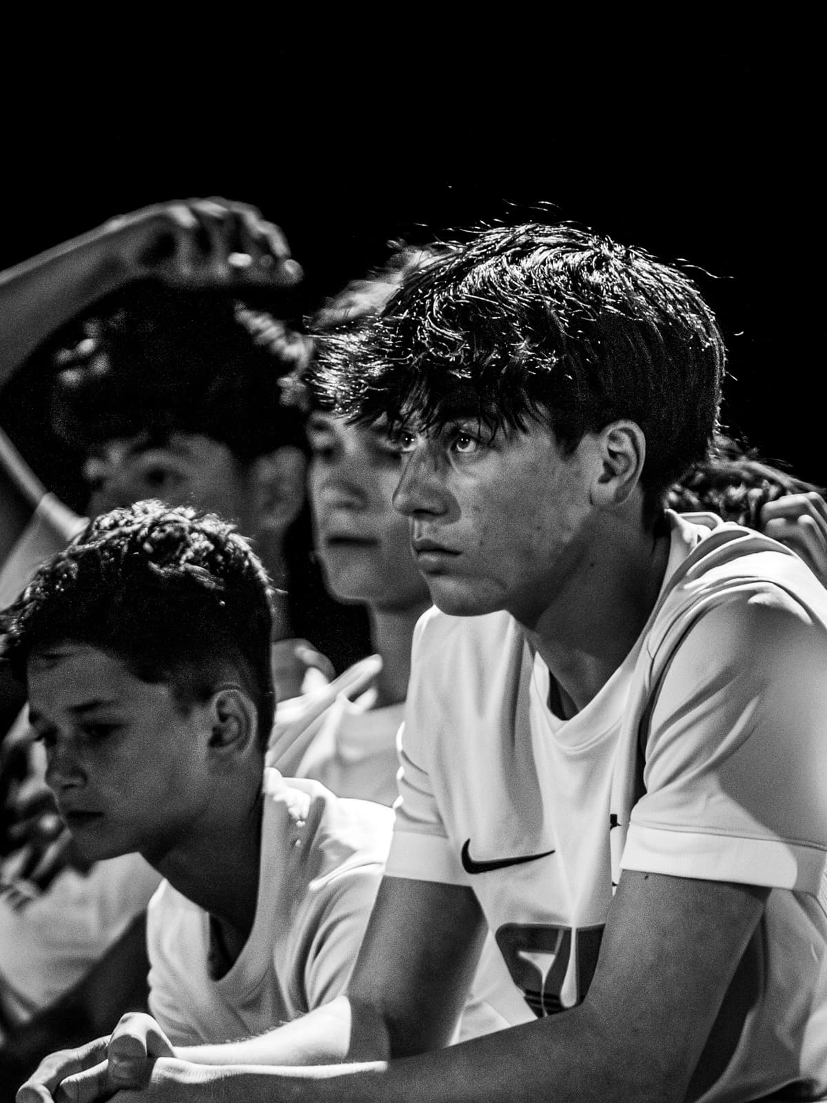 Black-and-white photograph of a row of young soccer players leaning on the bench rail, one looking up intently at the game.