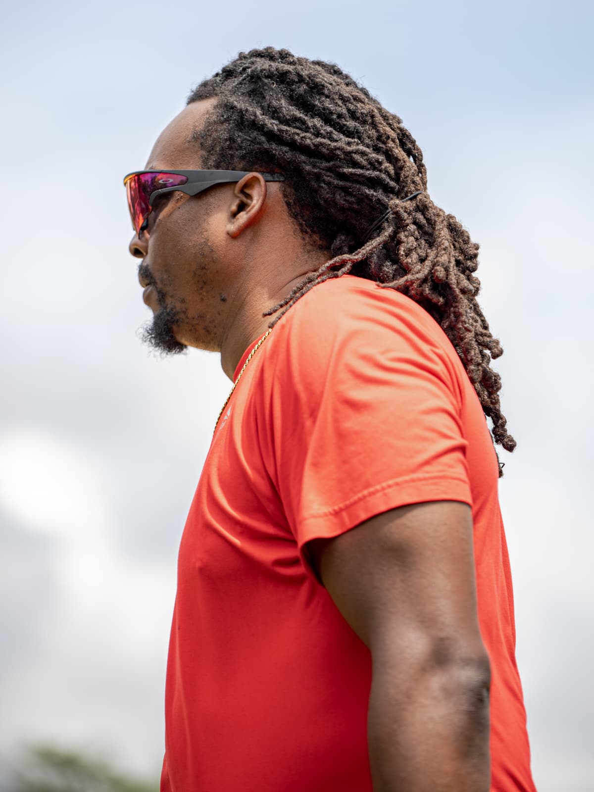 Profile portrait of a coach in an orange shirt and mirrored sunglasses watching from the sideline under an overcast sky.