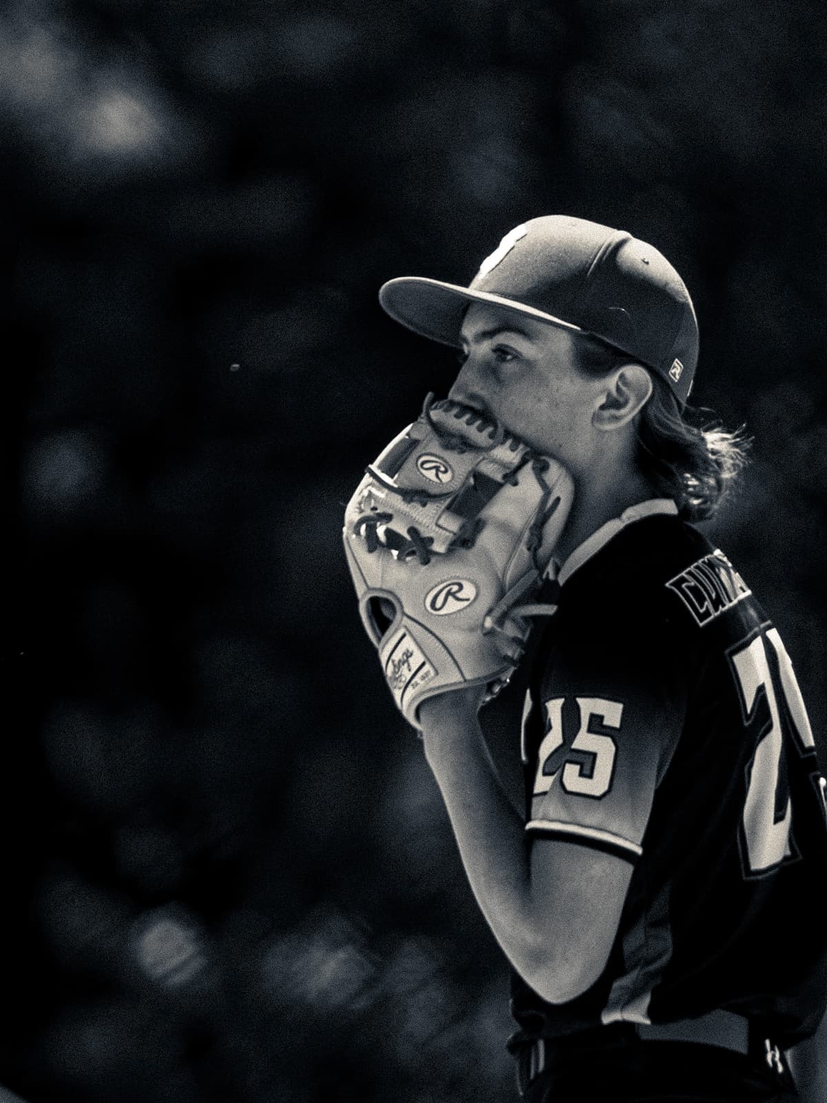 Black-and-white photograph of a young pitcher in a dark number 25 jersey holding his glove to his face as he looks in for the sign.
