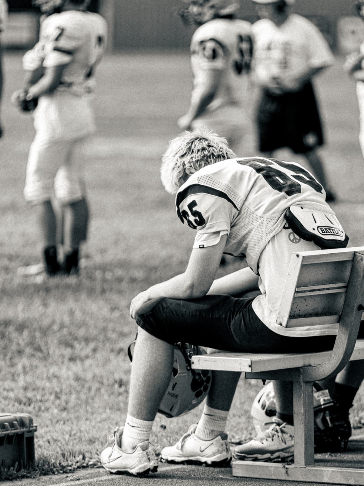 Black-and-white photograph of a youth football player in a number 65 jersey slumped forward on the sideline bench, head bowed.