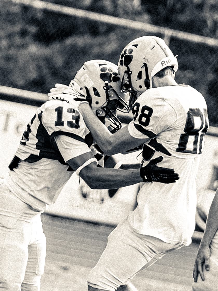 Sepia-toned photograph of two youth football players in white uniforms and paw-print helmets meeting helmet-to-helmet to celebrate.