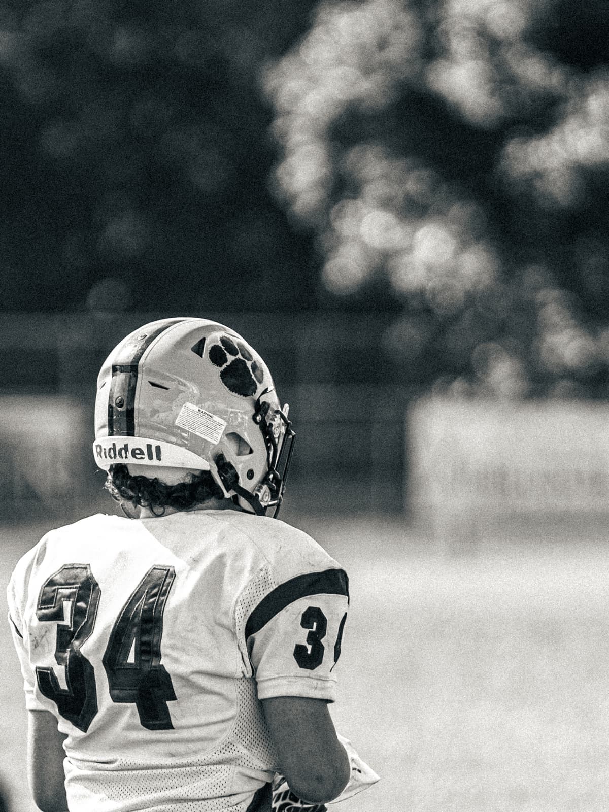 Black-and-white photograph of a youth football player in a number 34 jersey and paw-print helmet, seen from behind against a blurred tree line.