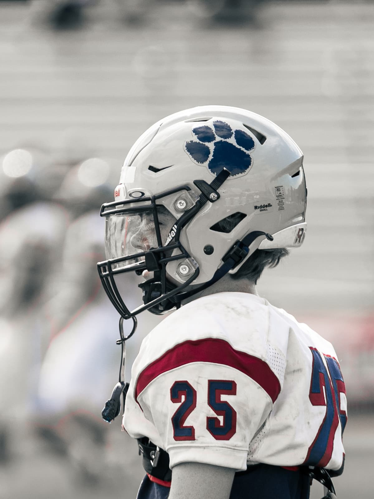 A youth football player in a white and red uniform and blue paw-print helmet looks down the line, seen in profile from behind.