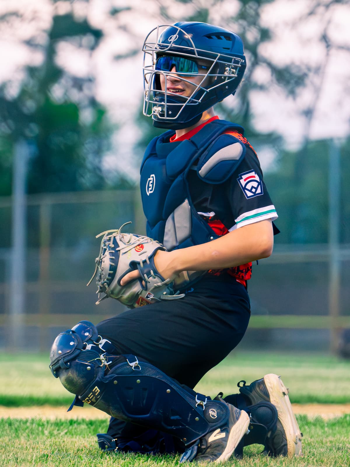 A Little League catcher in full navy gear kneels on the grass between innings, mask on and mitt resting across one knee.
