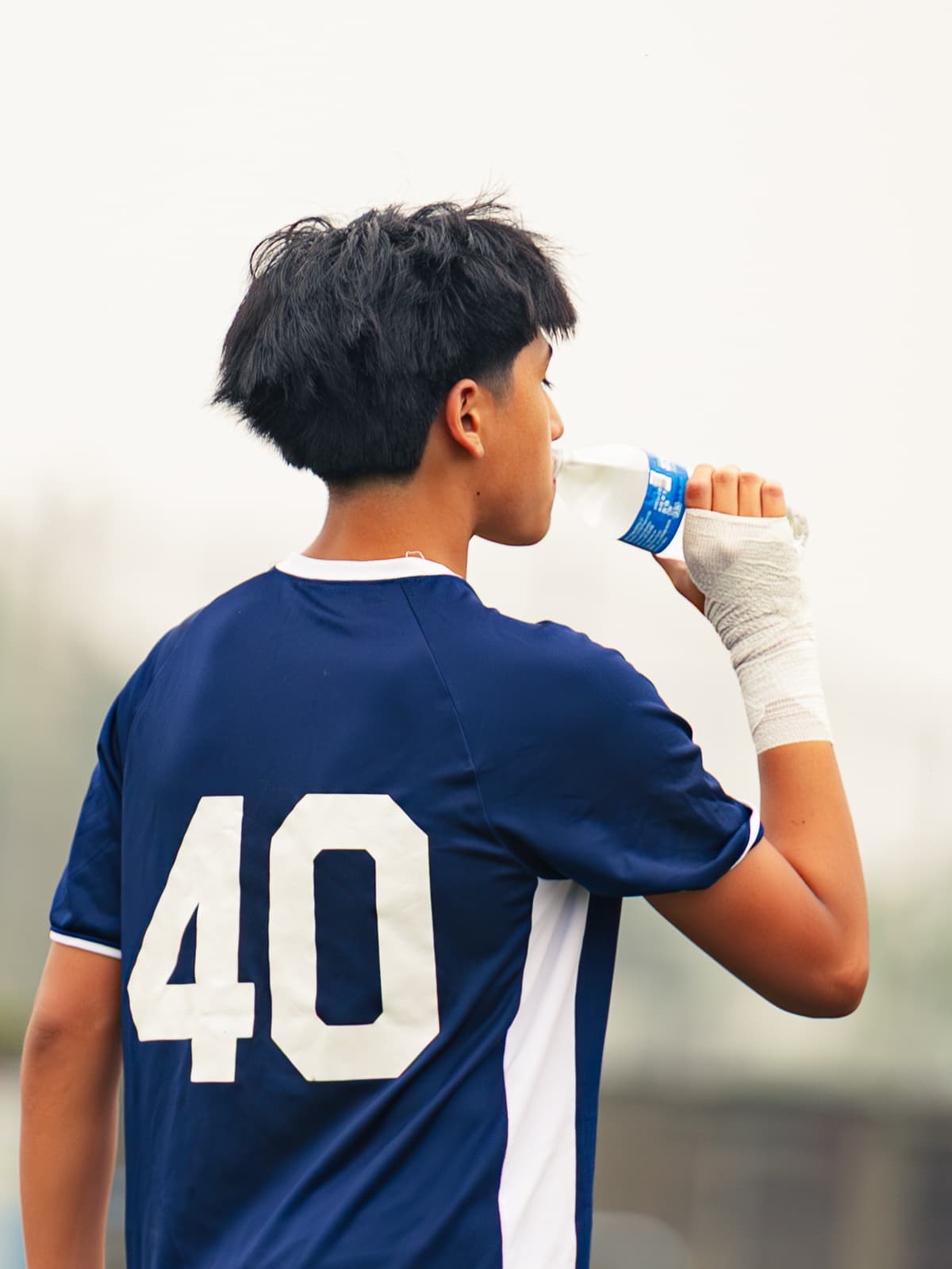 A soccer player in a navy number 40 kit drinks from a water bottle with a taped hand, seen from behind against a hazy sky.