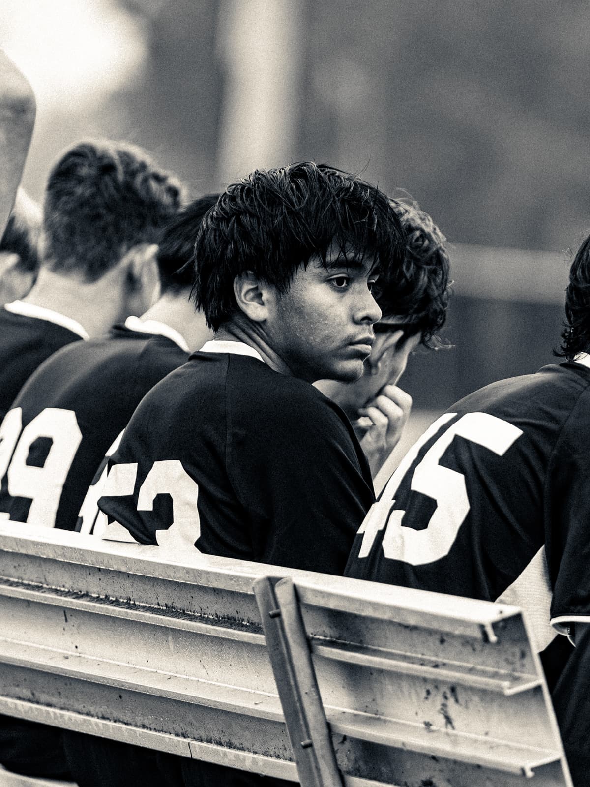Sepia-toned photograph of soccer players in warm-up jackets along the bench, a number 43 in the middle resting his chin on his hand.