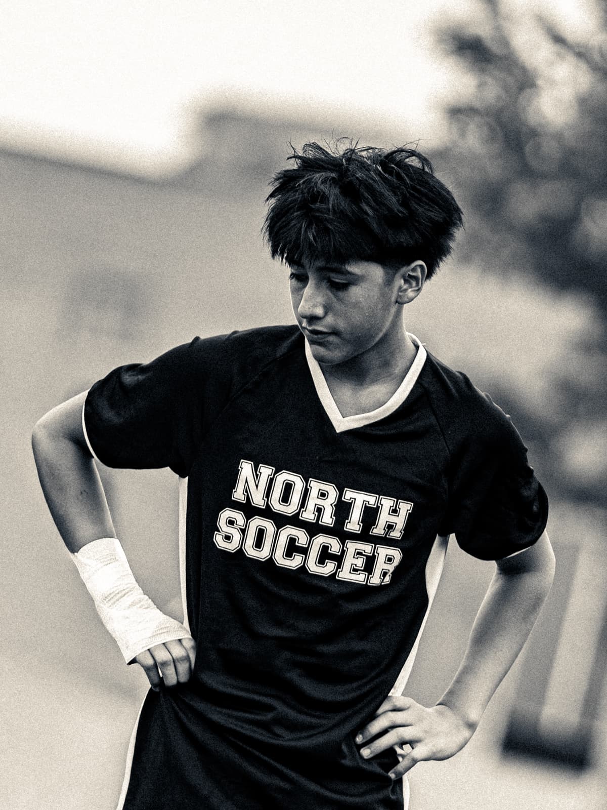 Black-and-white photograph of a soccer player in a North Soccer jersey standing with his hands on his hips and a taped wrist, catching his breath.