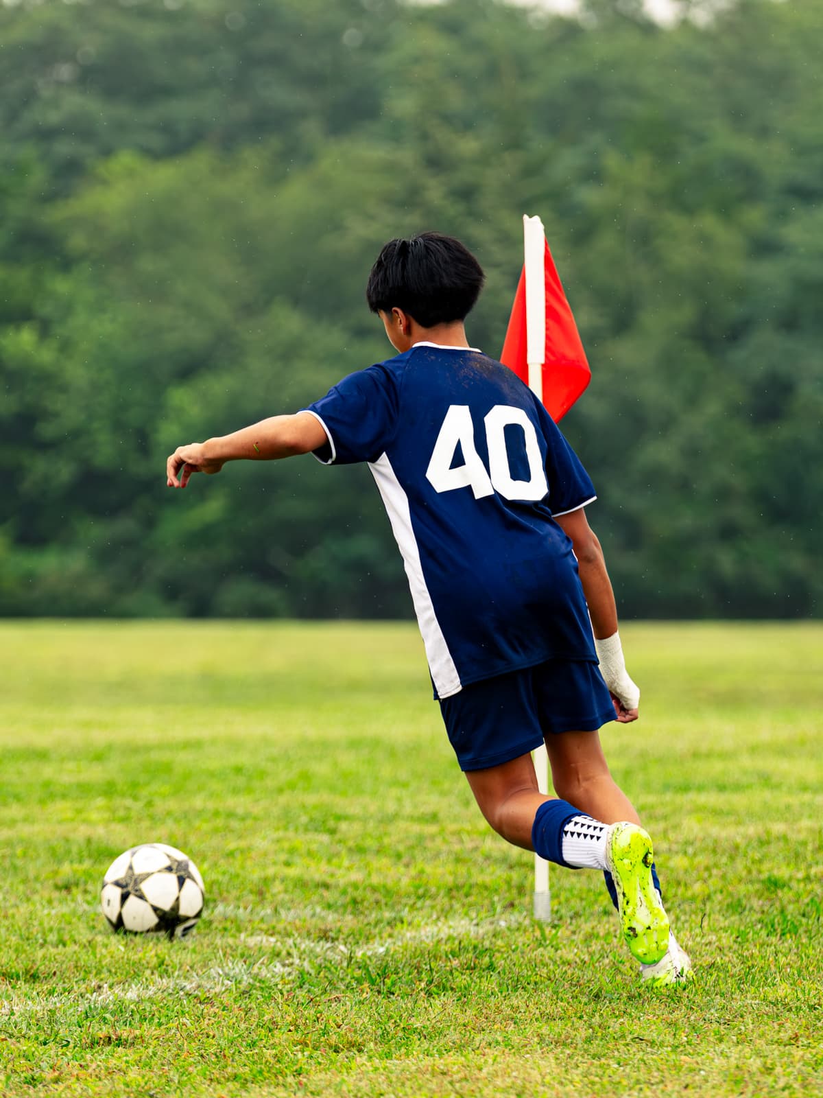 A soccer player in a navy number 40 kit runs onto the ball to take a corner beside the red corner flag, seen from behind.