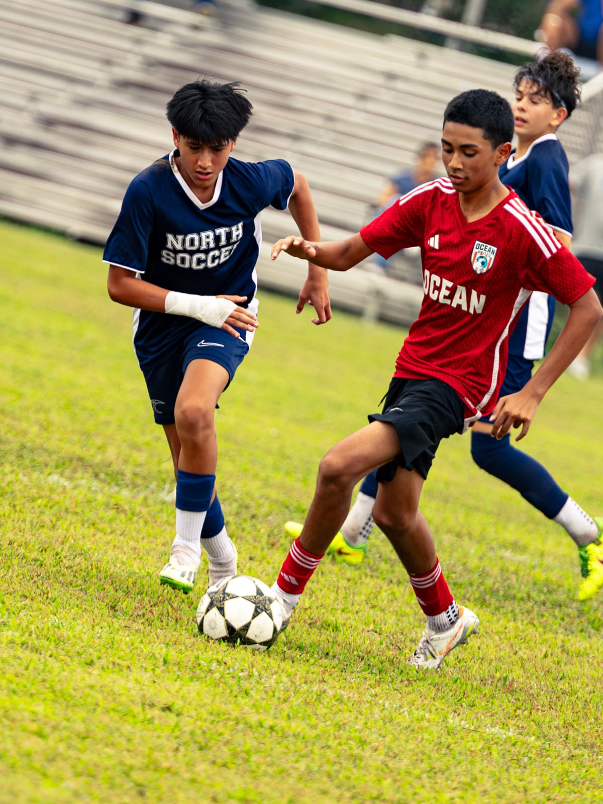 A soccer player in a navy North Soccer shirt and one in a red Ocean United shirt lean into each other for a loose ball at midfield.