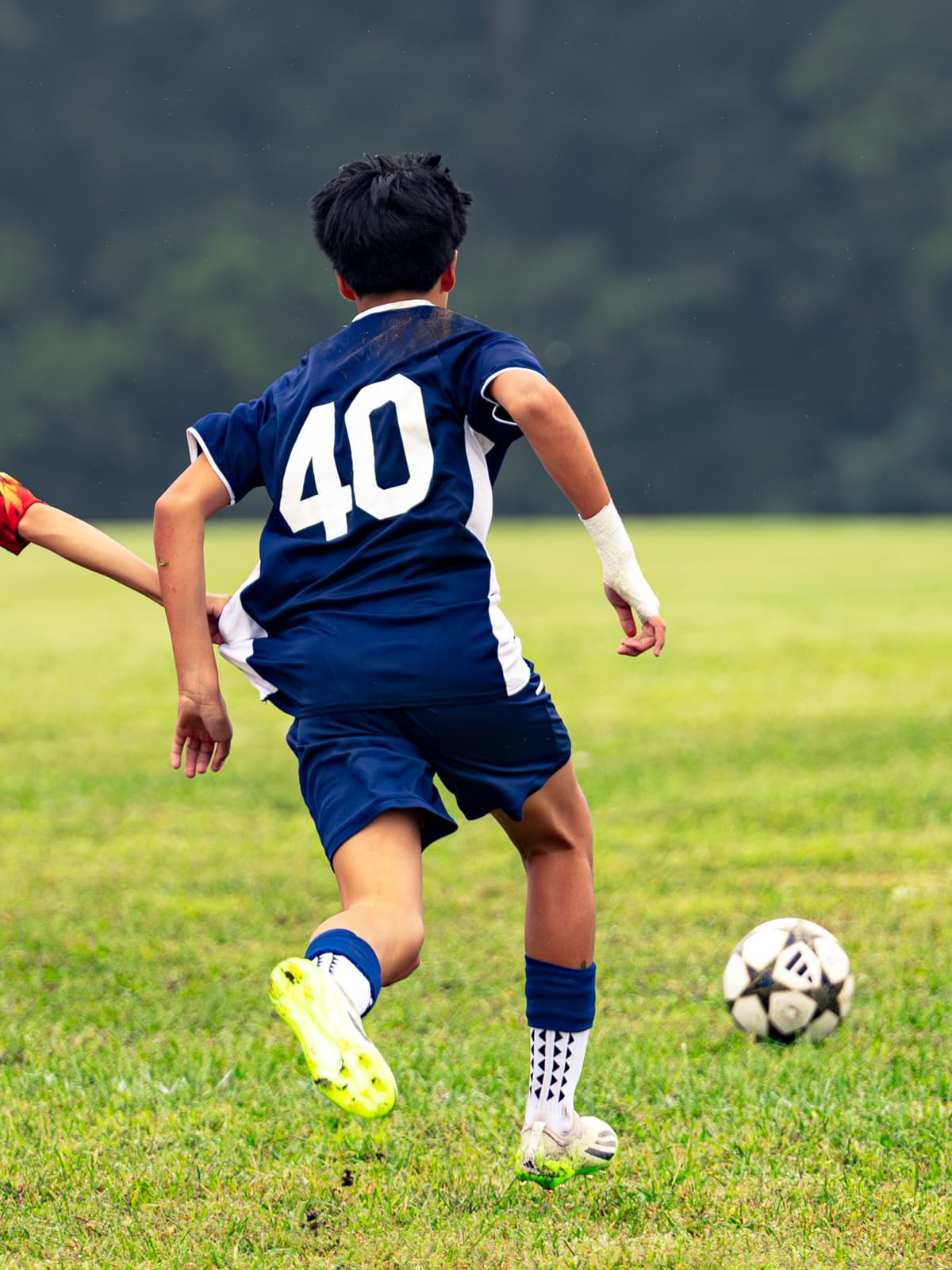 A soccer player in a navy number 40 kit strikes the ball with his laces at a full run, seen from behind as a defender lunges in.