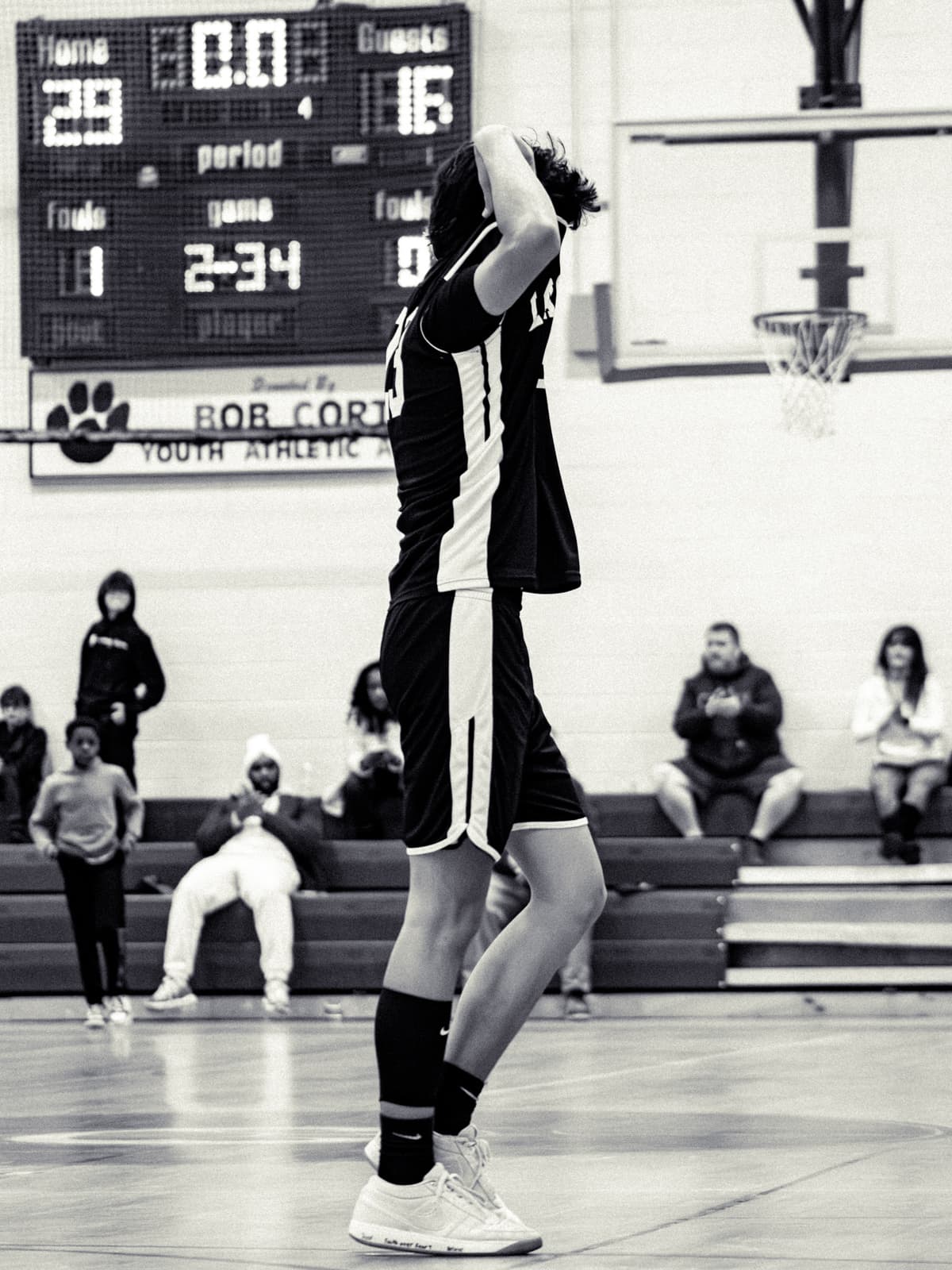 Black-and-white photograph of a youth basketball player at the free-throw line with both hands on his head, the gym scoreboard reading all zeros behind him.
