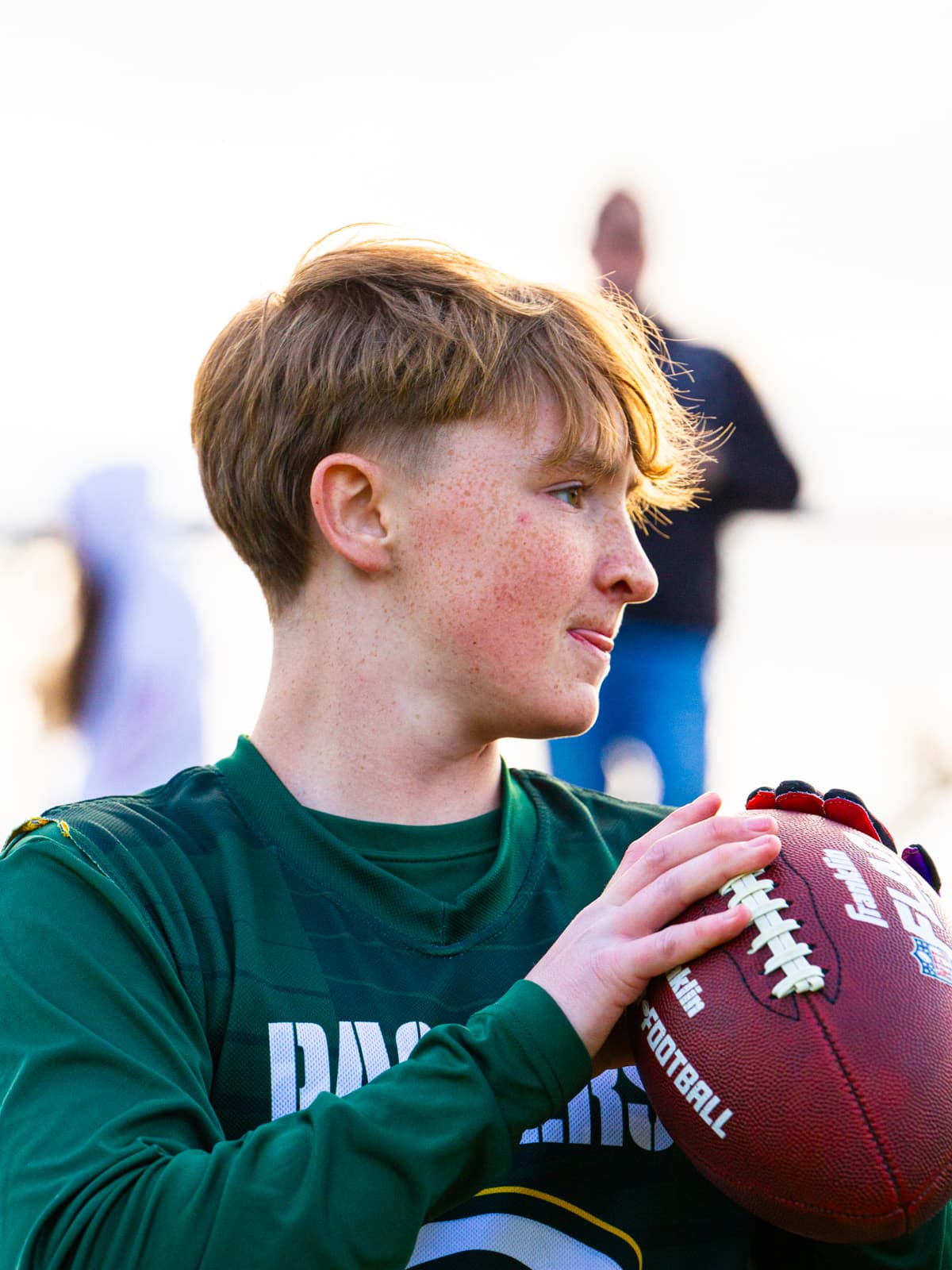 A young flag-football player in a green Packers jersey holds a football to his chest in golden light, looking out toward the field.