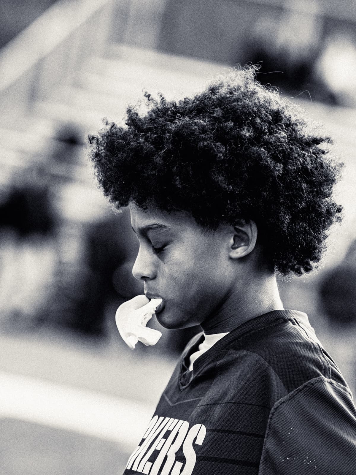 Black-and-white photograph of a young football player in a Panthers jersey with his mouthguard hanging from his lips, eyes down.