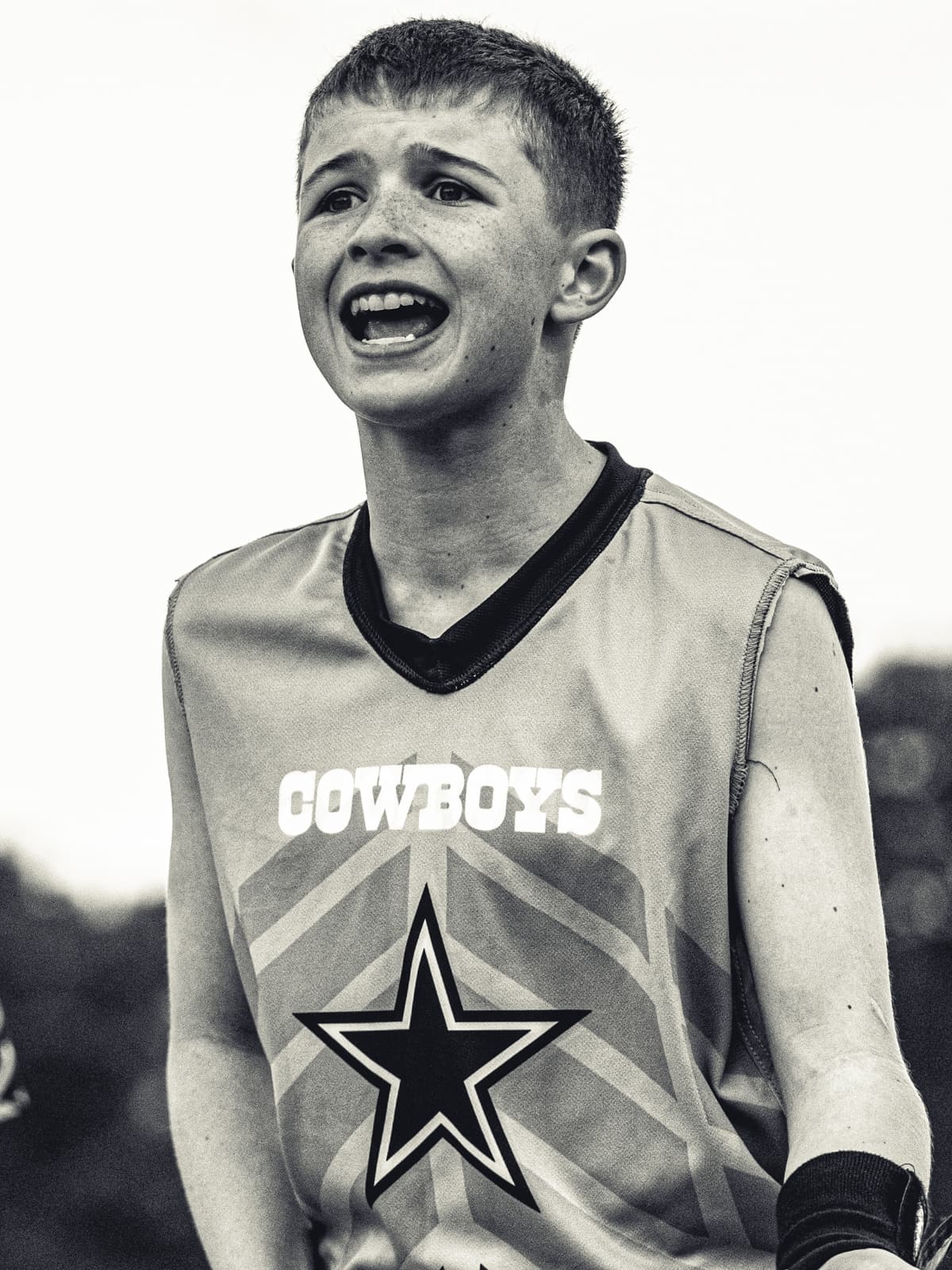 Black-and-white photograph of a young flag-football player in a Cowboys pinnie shouting toward the field, freckles catching the light.
